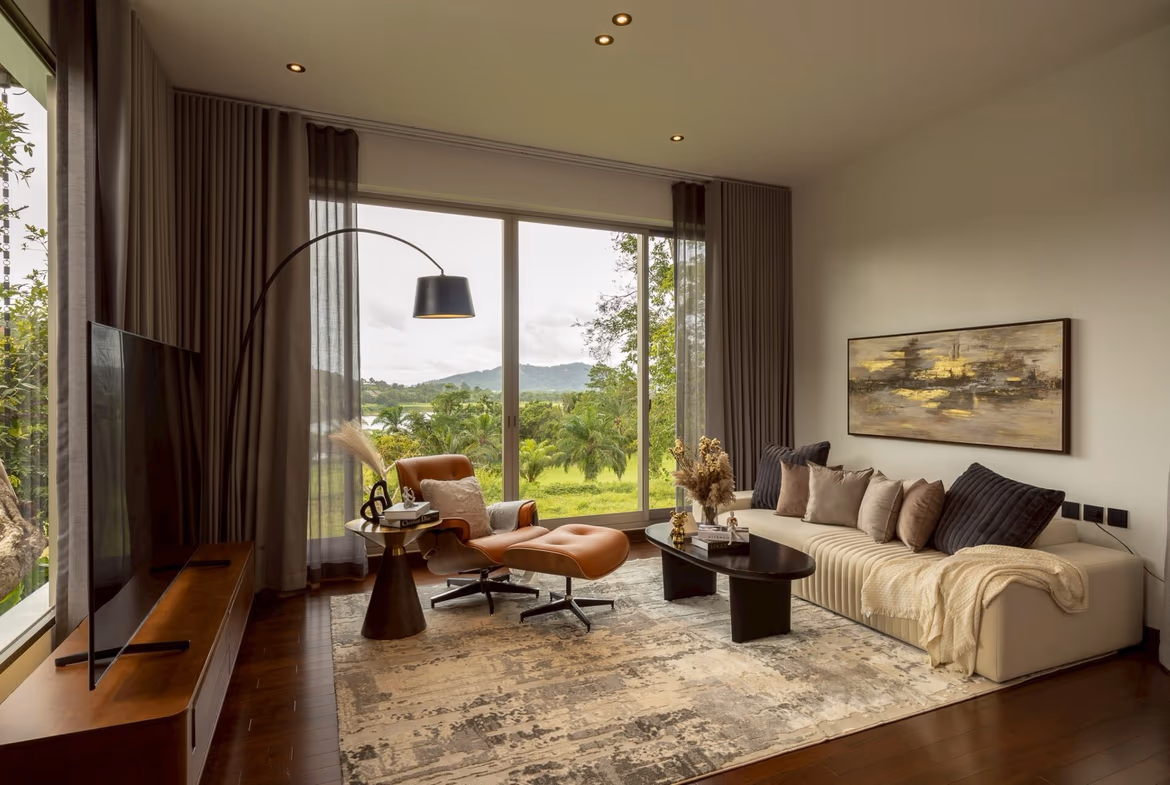 Bright living room with floor-to-ceiling windows and sheer gray curtains overlooking a green landscape; tan sofa and modern black coffee table.