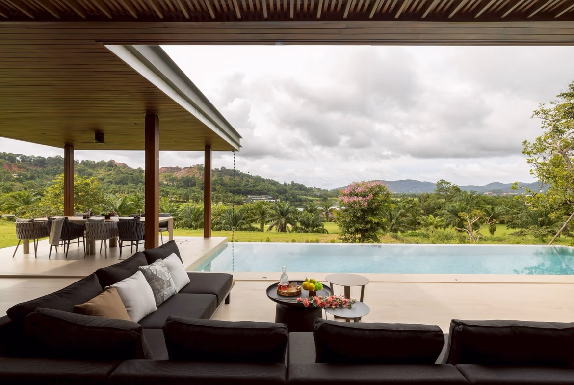 Outdoor living area with a black sectional sofa and round coffee tables, pool in foreground, covered patio, and lush green landscape with hills in the distance under a cloudy sky.