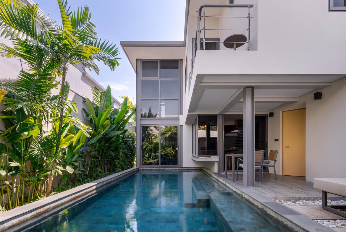 Modern two-story villa with a rectangular pool, lush tropical plants along the left, and an outdoor dining area under a covered patio.