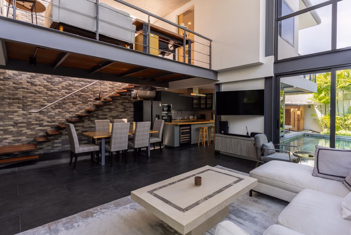 Open-plan living area with dining table, kitchen, and a wooden staircase beneath a mezzanine balcony.