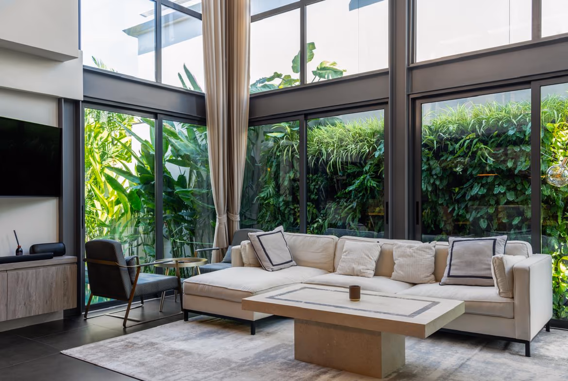 Bright contemporary living room with floor-to-ceiling glass walls, beige sectional sofa, and lush tropical plants outside.