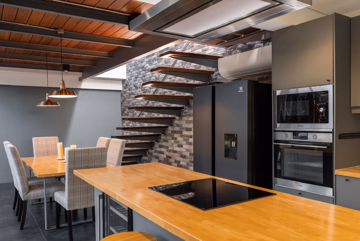 Open-concept kitchen with a wooden island, black induction cooktop, and stainless steel fridge and ovens beside a floating wooden staircase against a brick wall.