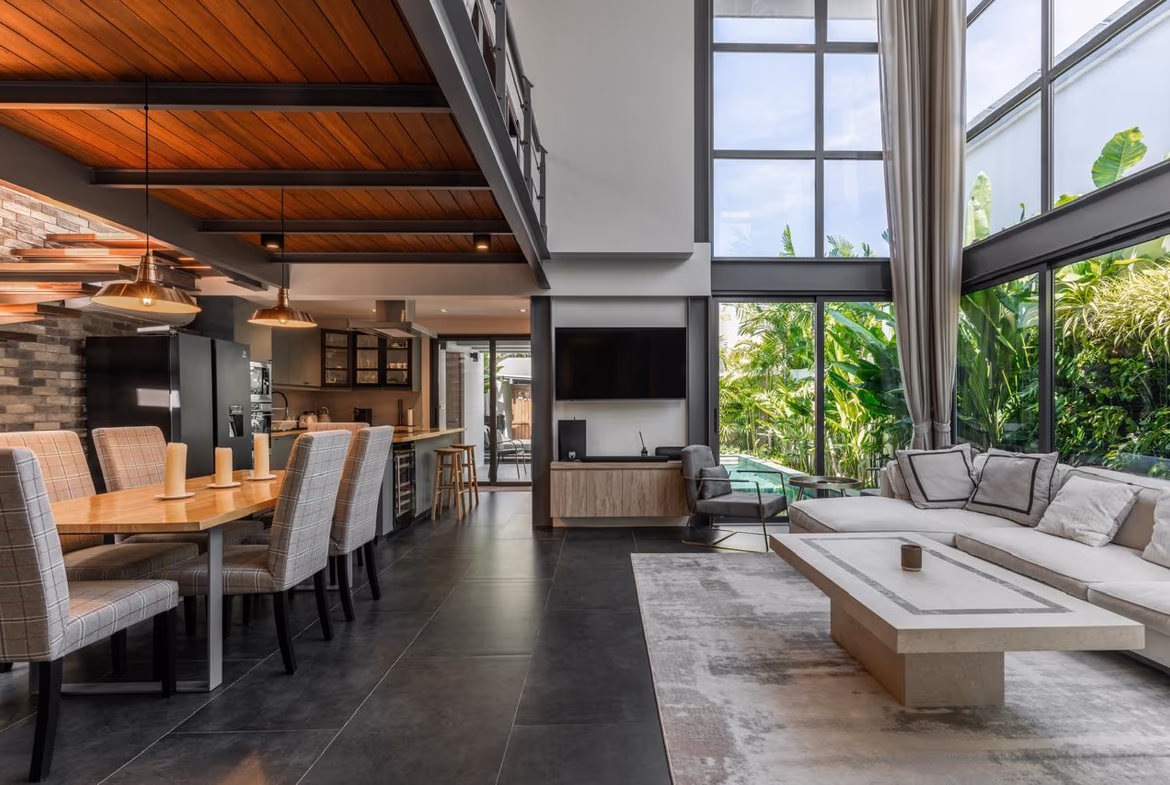 Open-plan living and dining area with a long wood dining table, upholstered chairs, a modern kitchen, and seating by large windows.