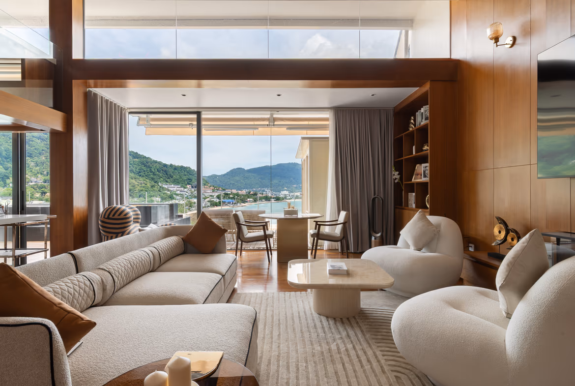 Luxurious living room with cream sectional sofas, curved chairs, and wood-paneled walls framing a glass door to an outdoor balcony overlooking a coastal town and green hills.
