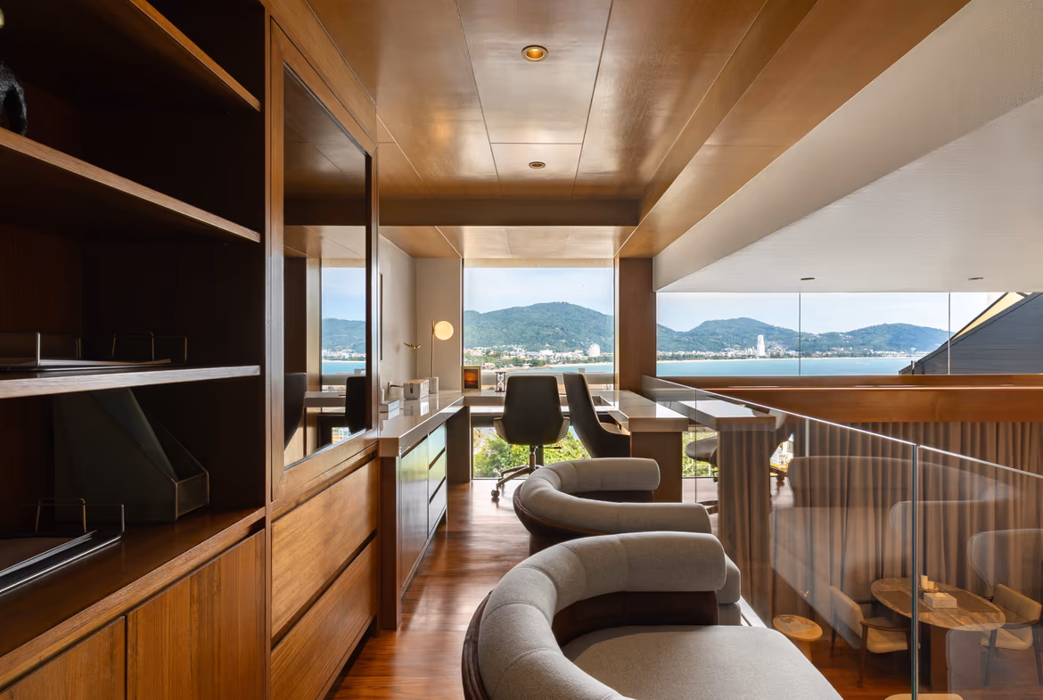Luxurious wooden office with curved gray chairs and a glass railing, overlooking a bay and mountains through large windows.