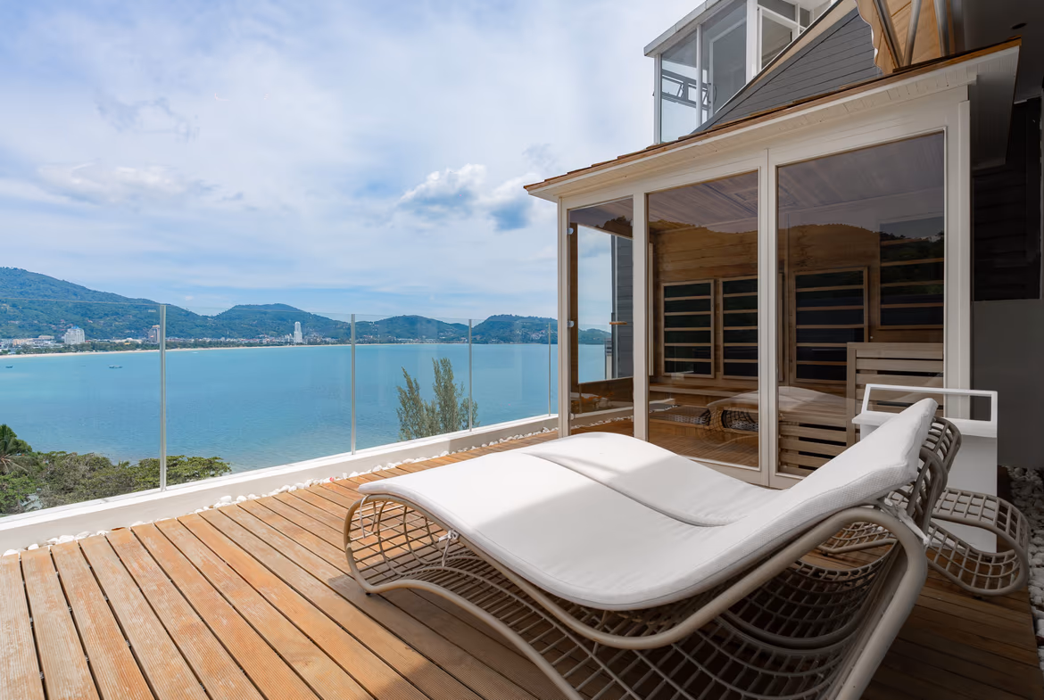 Sunlit wooden terrace with white lounge chairs overlooking calm blue ocean and distant hills through a glass railing