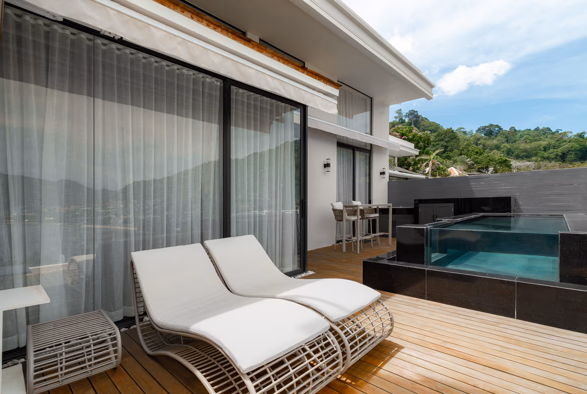 Outdoor poolside patio with white lounge chairs on a wooden deck beside glass sliding doors and sheer curtains behind them