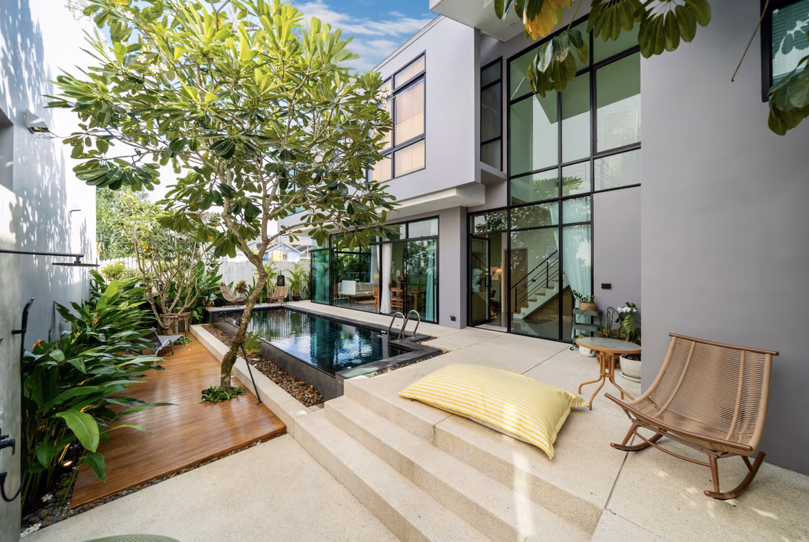 Modern two-story home courtyard with a narrow pool, glass walls, and lounge seating amid tropical plants.