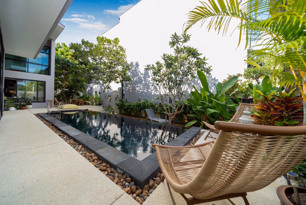 Modern courtyard with a rectangular pool, tan deck, and wicker lounge chairs amid tropical plants by a white building.