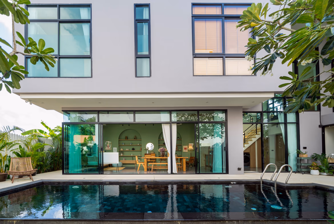 Modern two-story home with large glass sliding doors, pool in foreground, and tropical plants on sides.