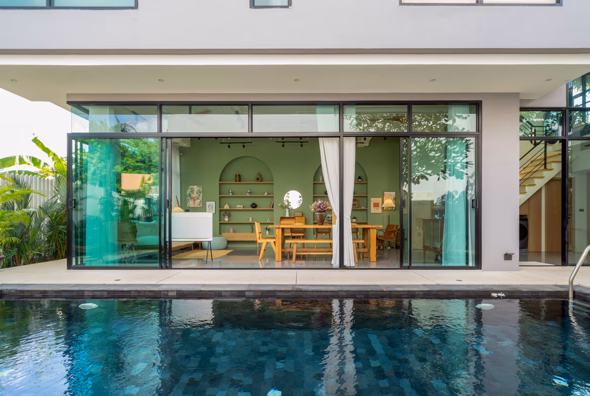 Open-plan living room with a green wall and wooden dining table seen through large glass sliding doors by a poolside patio.
