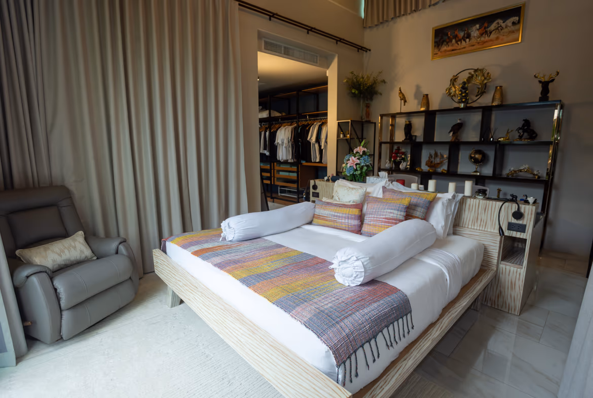 Bedroom with a low wooden platform bed, colorful throw, and rolled white bolster pillows in front of a display shelf.