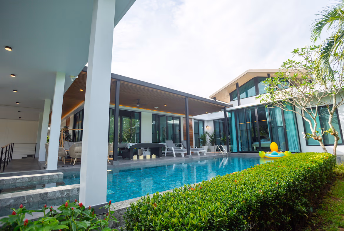 Resort-style villa with a rectangular pool, outdoor seating under a covered patio, and tropical hedges along the edge.