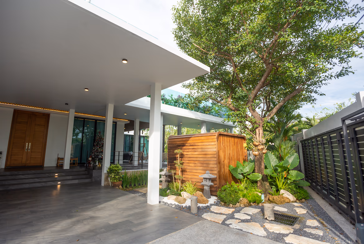 Modern covered patio with white columns, dark tile flooring, and a wooden door at the left; garden area with rocks and greenery in the center-right, and a curved wooden shed behind.