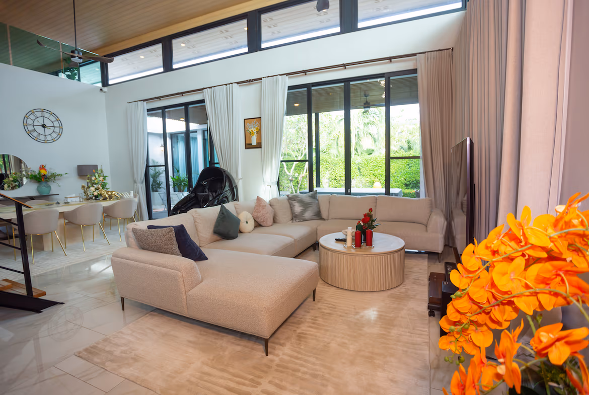 Bright open-plan living room with a beige sectional, round coffee table, and orange flowers in the foreground; large windows with cream curtains overlook a garden area.