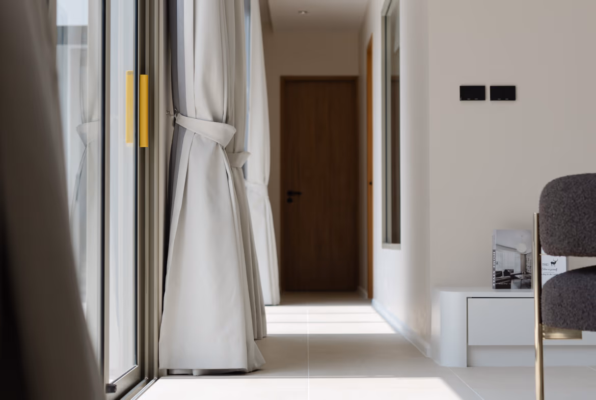 Bright modern hallway with floor-to-ceiling windows and tied-back curtains on the left, leading to a wooden door at the end.