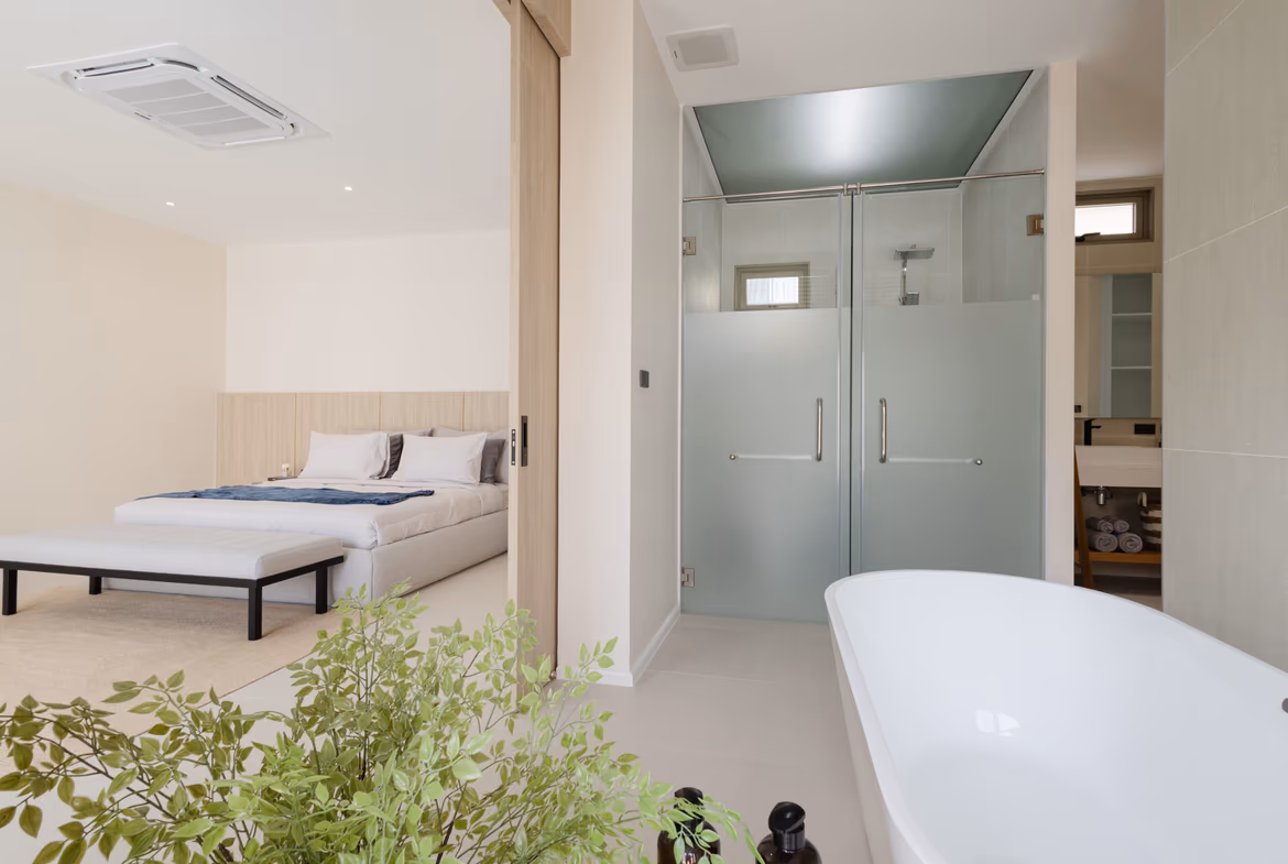 Open-plan hotel suite showing a bed area adjacent to a glass-walled bathroom with a frosted lower panel and a freestanding bathtub in the foreground, plus green plants in front.