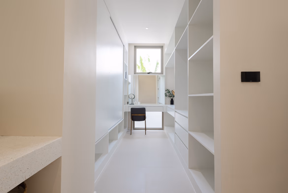 Bright walk-in closet with white built-in shelves, a desk, and a window at the far end letting in light.