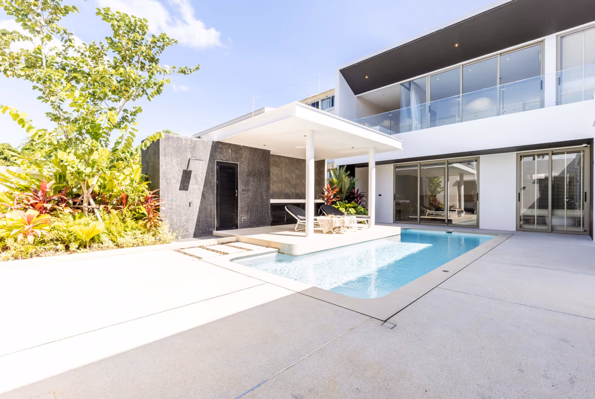 Modern house with a rectangular swimming pool, white patio, and glass balcony railing; lounge chairs under a shaded canopy.