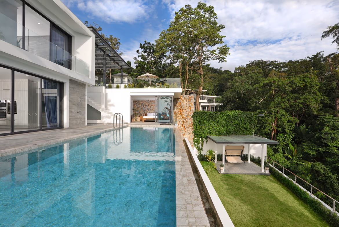 Modern white villa with large glass walls and a rectangular swimming pool, overlooking a green hillside.
