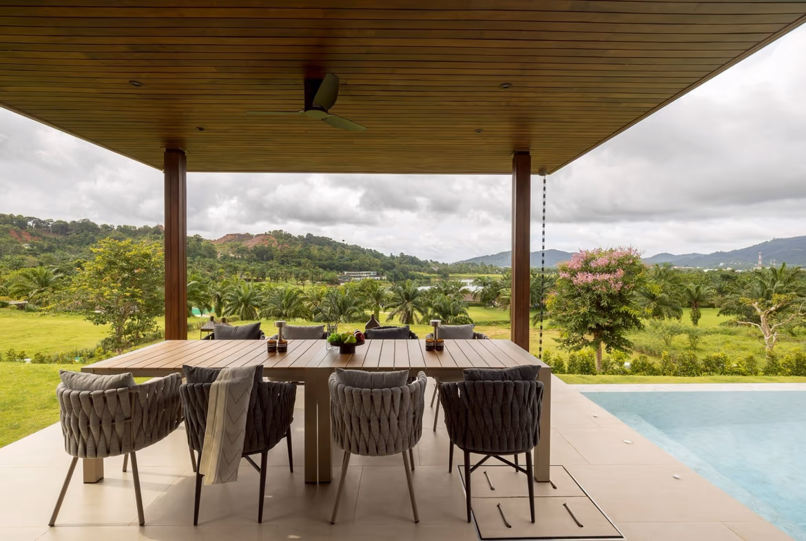 Outdoor covered dining area with a long wooden table and woven chairs, overlooking a lush landscape of trees and hills by a poolside patio.