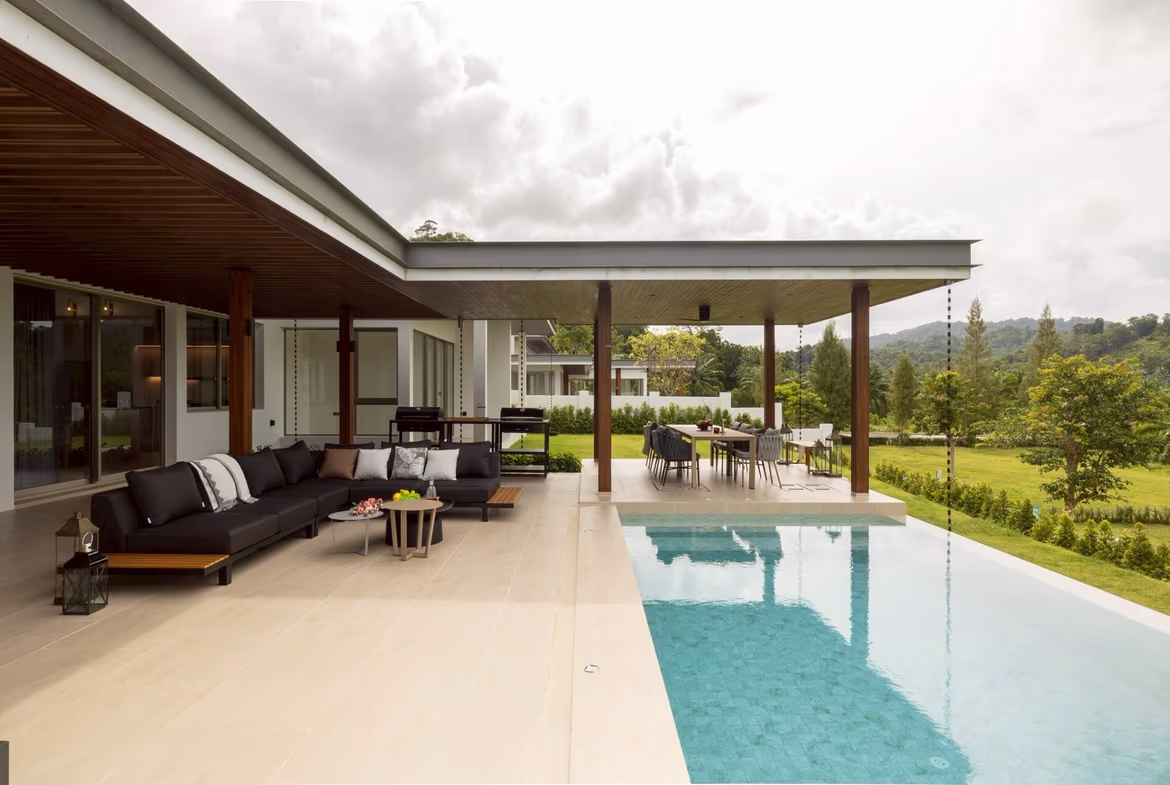 Modern outdoor living area with beige tiled patio, black sectional sofa, and a rectangular pool full of crystal water.