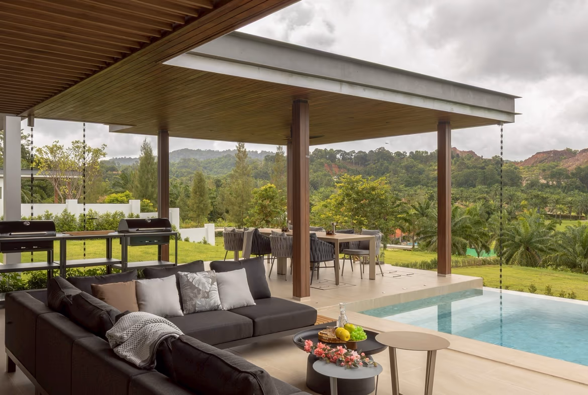Outdoor covered patio with a dark sectional sofa, coffee tables, and a rectangular pool, overlooking lush green hills.