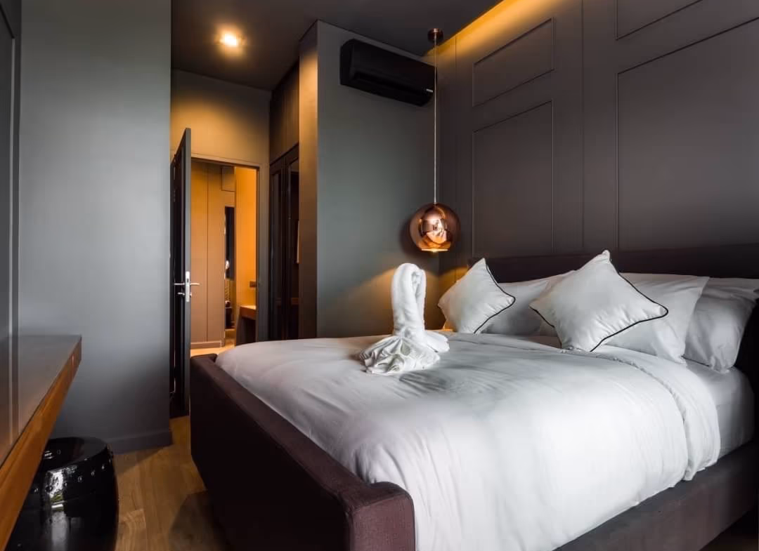 Modern hotel bedroom with a large white bed, gray walls, and a copper pendant light over the side; towel folded into a swan on the blanket.