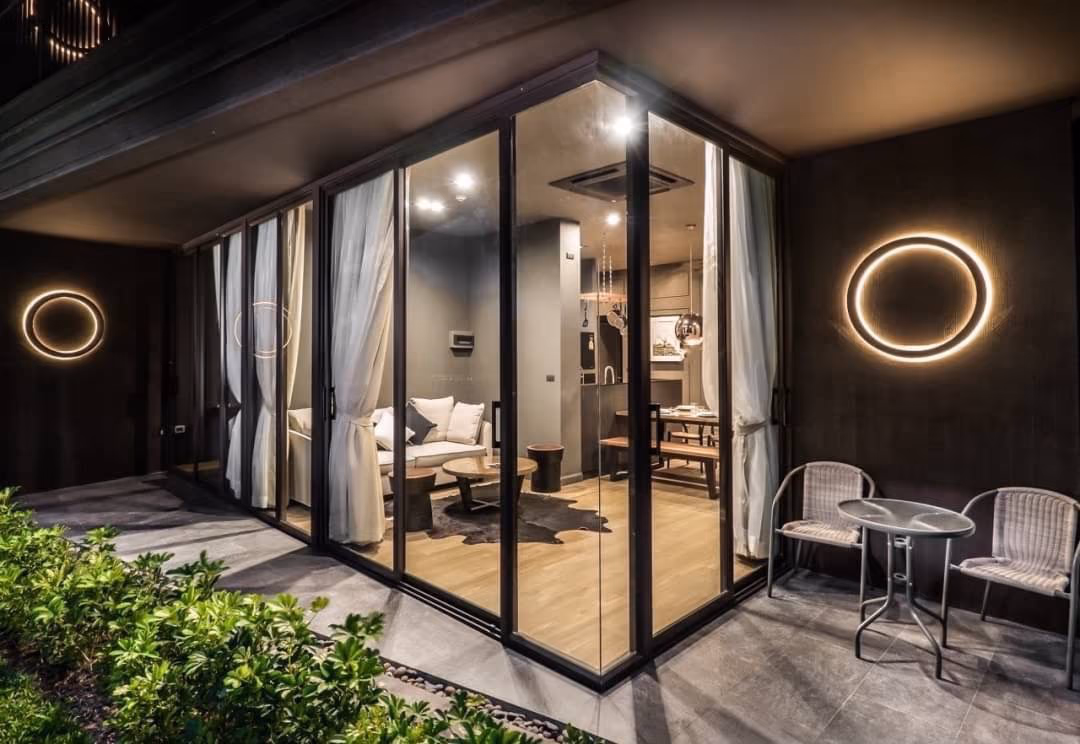 Contemporary glass-walled living room at night, featuring a sectional sofa, wooden coffee tables, and a visible kitchen, accented by circular wall lights outside.