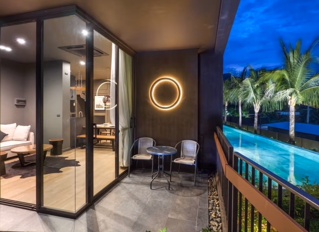 Hotel balcony at dusk with two chairs and a small table, circular wall light, and a pool with palm trees.