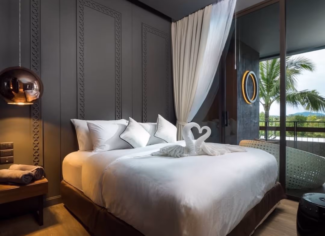 Luxurious hotel room with a large white bed, decorative gray paneled wall, and a balcony view with palm trees outside the glass door; bed has towel swans on top.