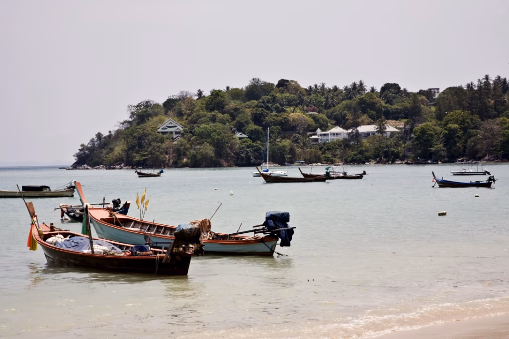 Small colorful fishing boats anchored near a light turquoise shore with a forested island and houses in the background