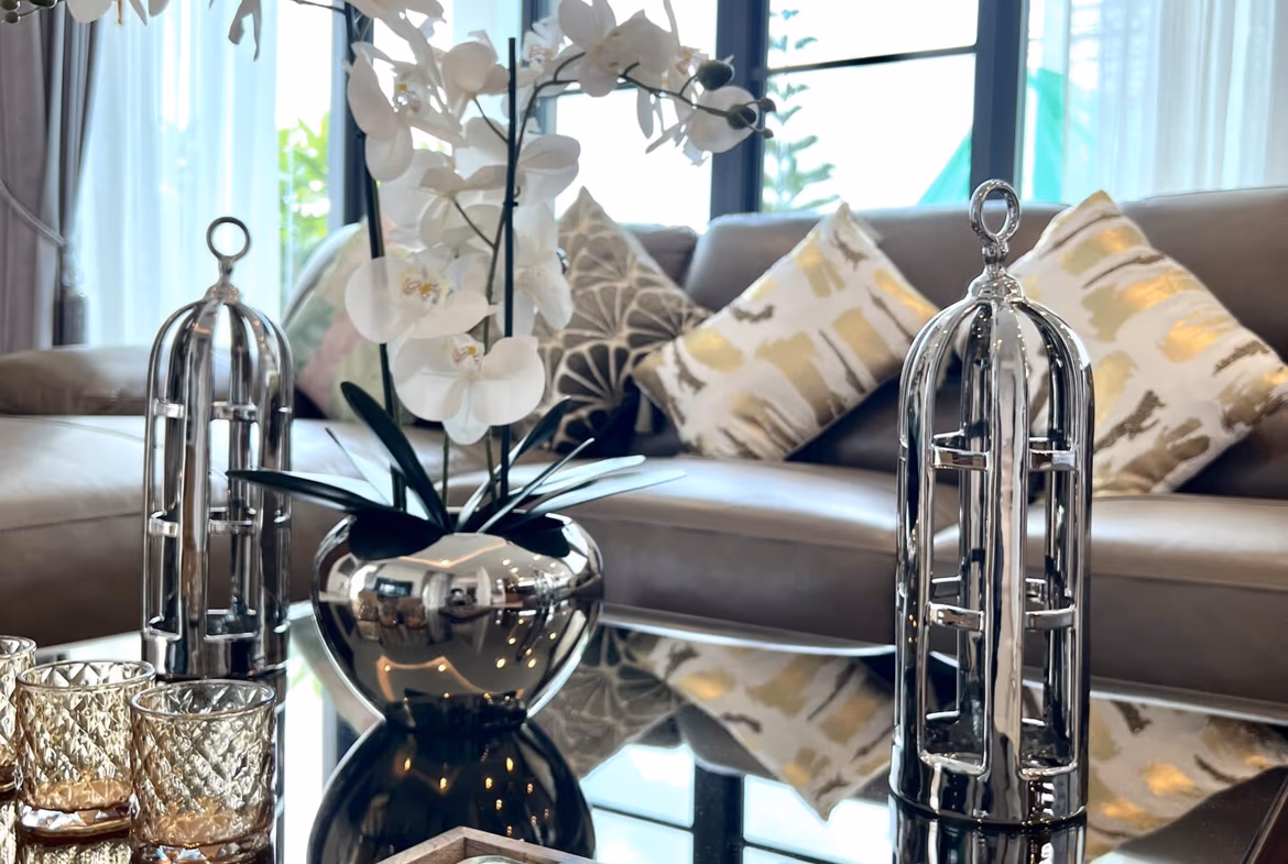 Modern living room centerpiece with white orchid in a shiny silver vase on a glass coffee table, reflective surfaces and patterned cushions in the background.