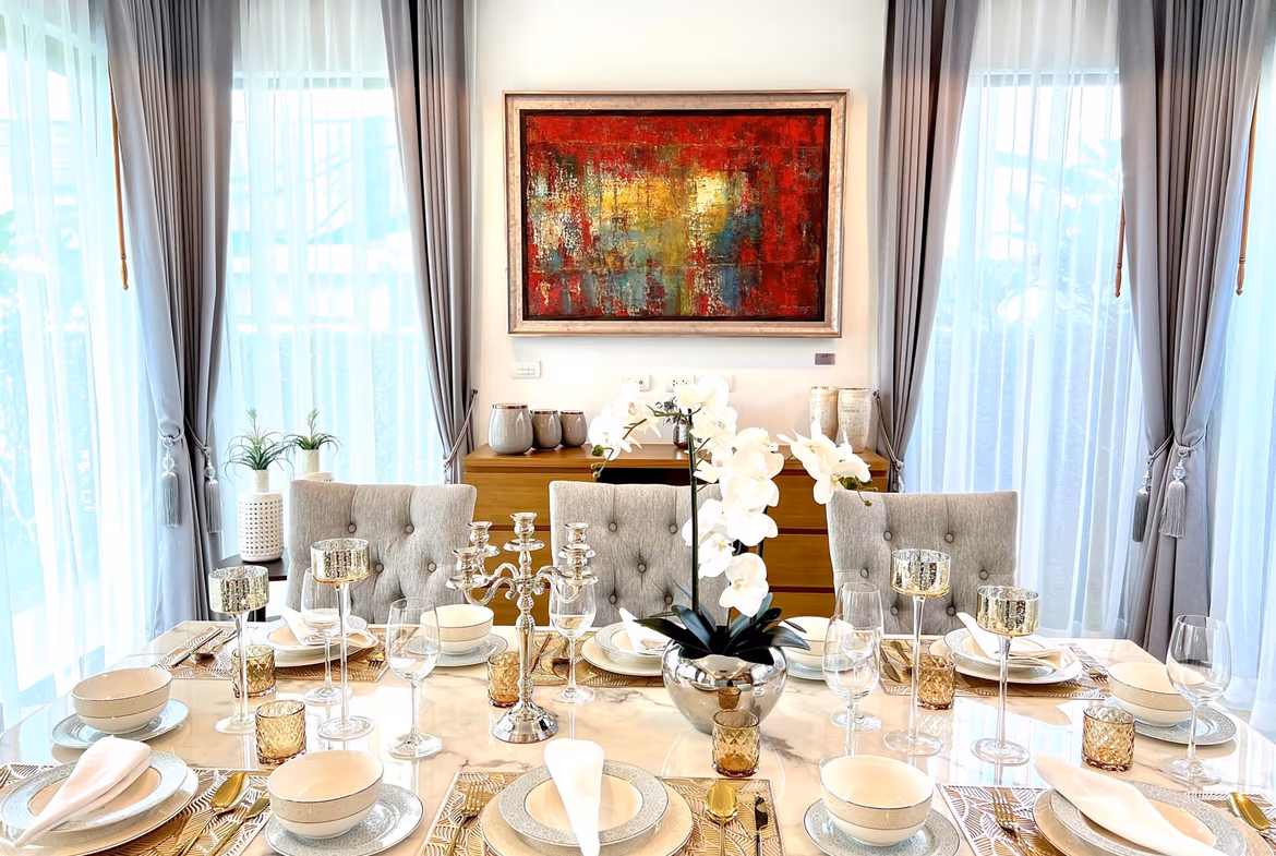 Elegant dining room with a long marble table set for a formal dinner, white napkins, fine china, crystal glassware, and gold-toned candlesticks, framed by gray curtains and a bold abstract painting on the wall.