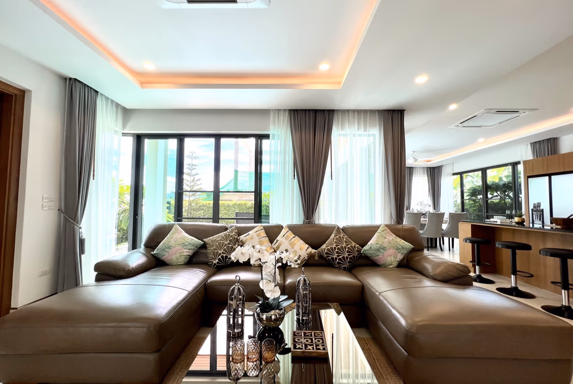 Open-plan living room with a brown L-shaped sectional, decorative cushions, and a glass coffee table near large sliding doors with sheer gray curtains.