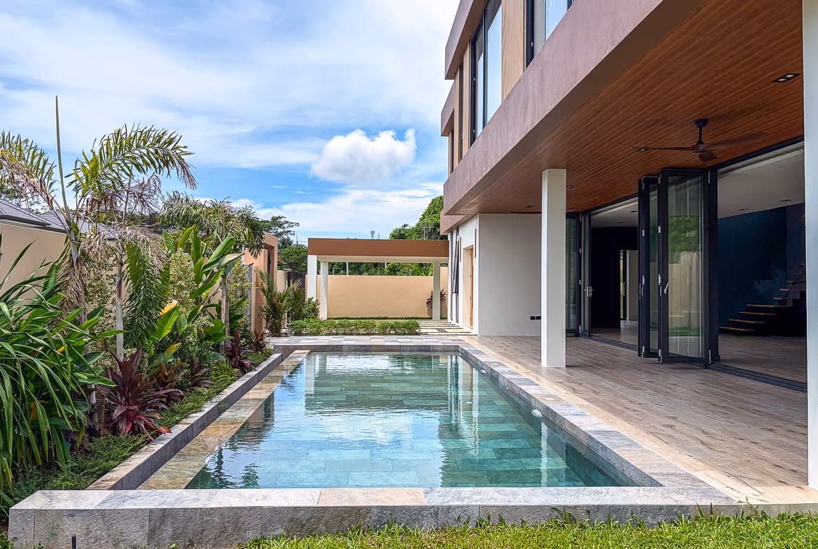 Rectangular pool beside a modern house with a wood-paneled ceiling, glass doors, and lush tropical plants along the left edge.