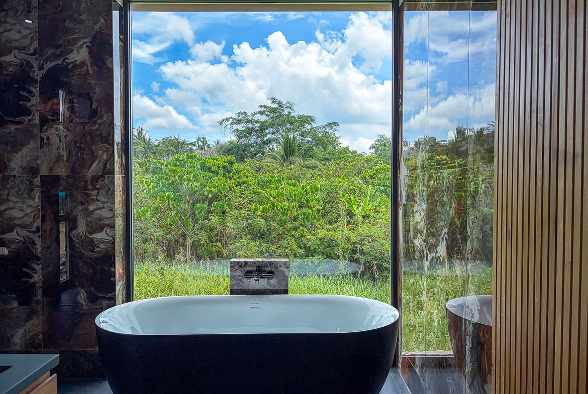 Freestanding black bathtub facing a large window with a lush green garden and blue sky outside, marble walls on the left and wooden paneling on the right.