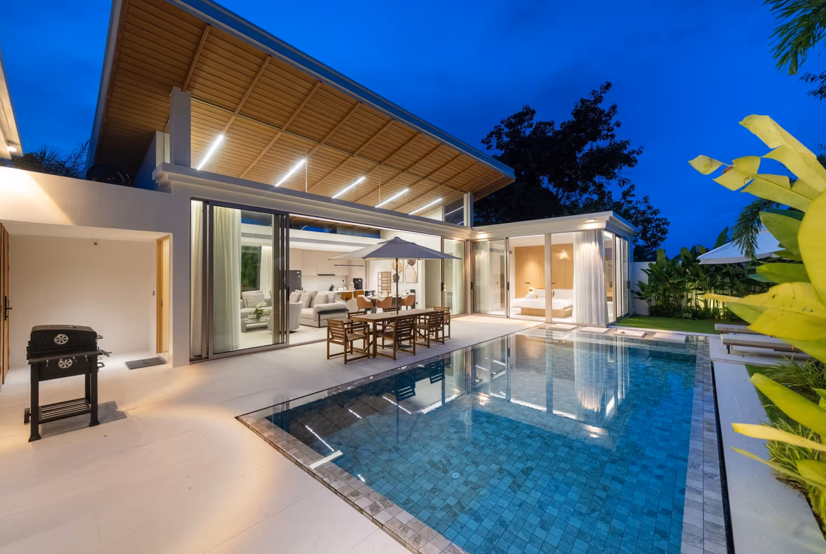 Modern house with an illuminated wooden pergola over a glass-walled living area, outdoor dining by a blue-tiled pool at dusk