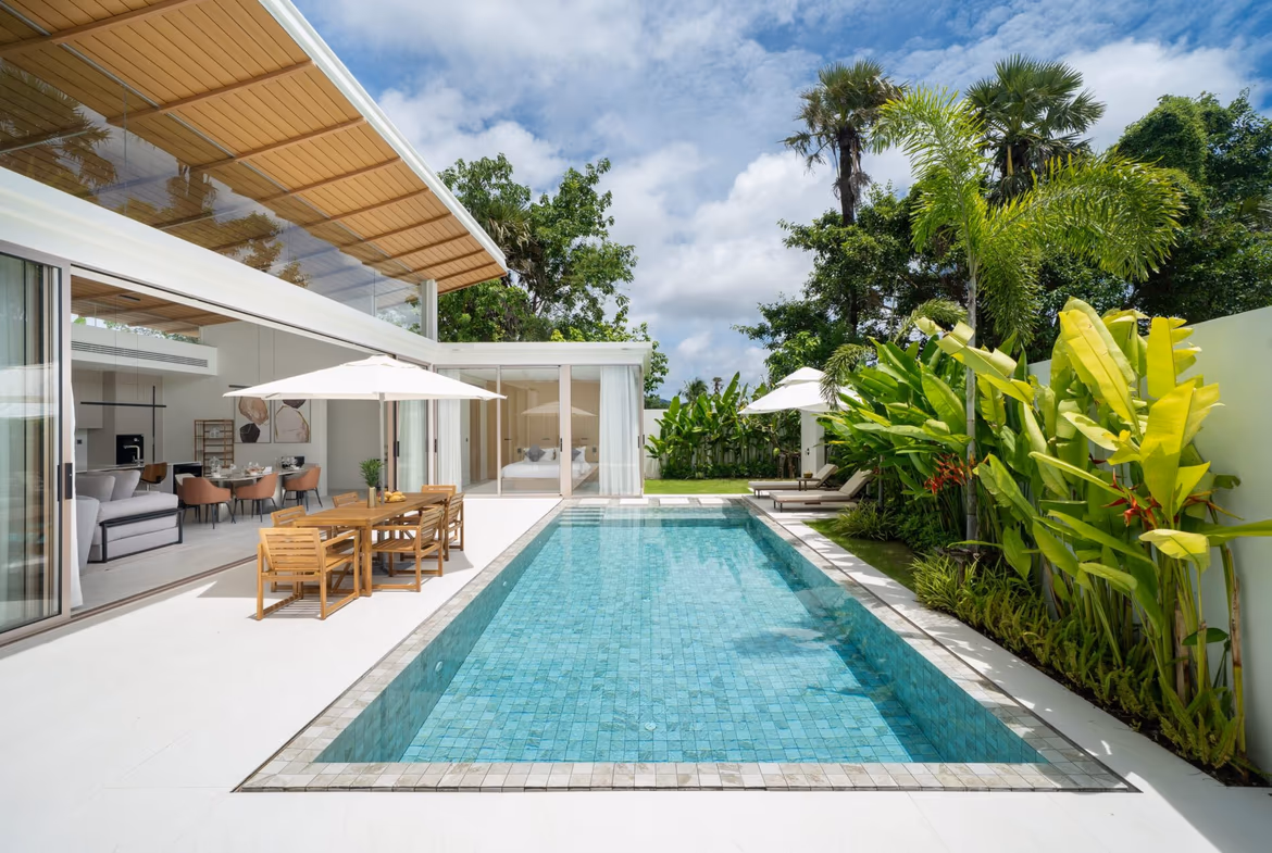 Modern backyard with a rectangular turquoise pool, glass-walled living area, wooden dining table under umbrellas, and tropical plants.