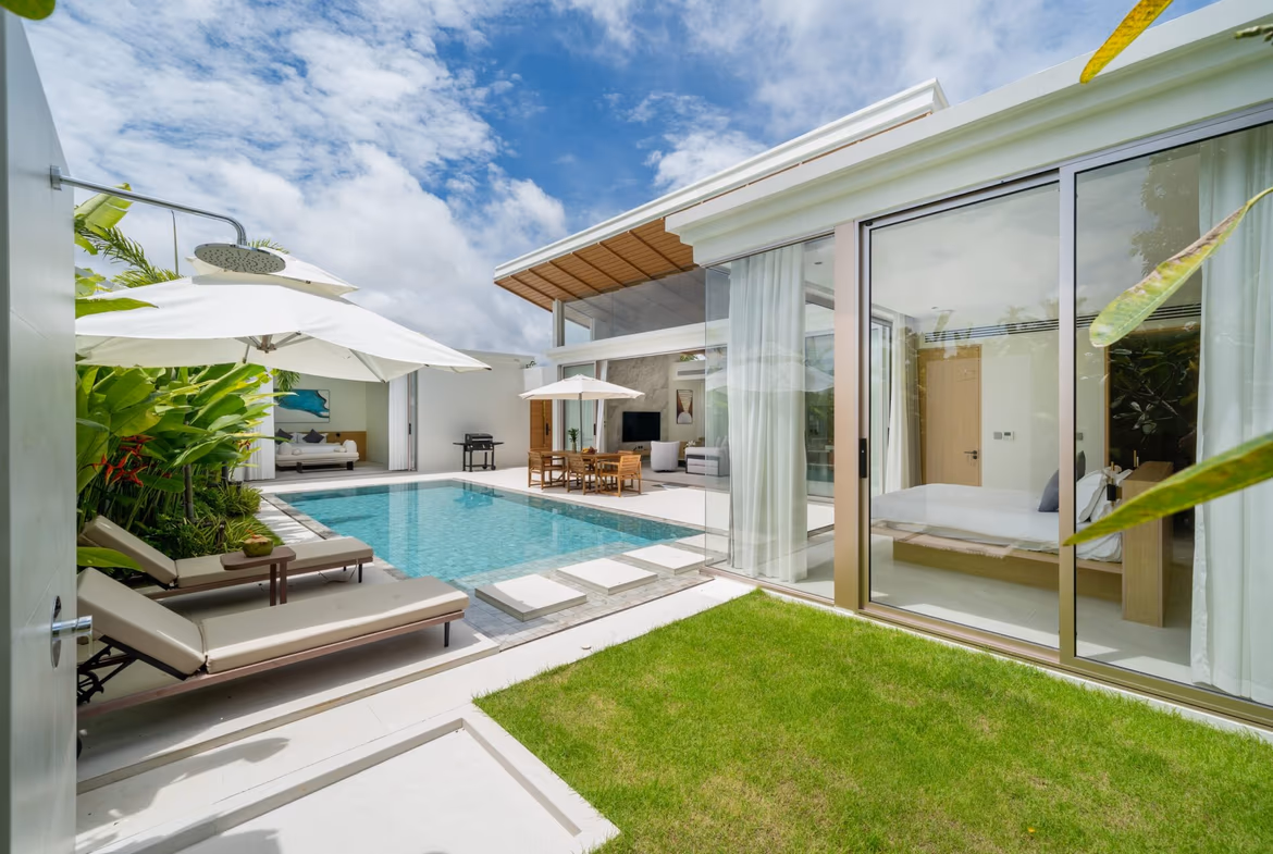 Modern villa backyard with a rectangular pool, white loungers, and umbrellas beside a glass-walled living area