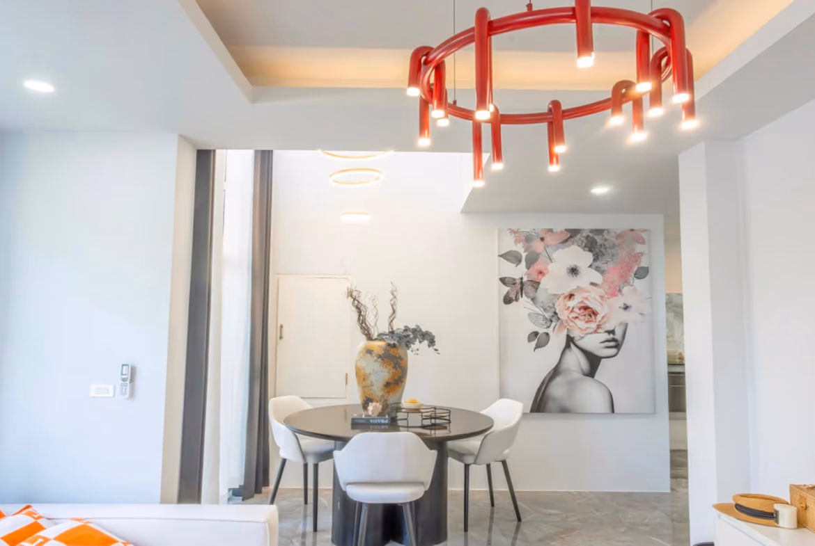 Modern dining area with a round table and white chairs, a bold red circular chandelier overhead, and a floral-art portrait on the wall.