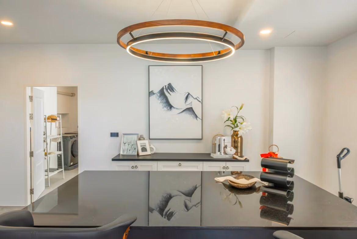 Modern kitchen with a glossy black island, circular wooden-and-light pendant overhead, and a mountain art print behind a white cabinet run.